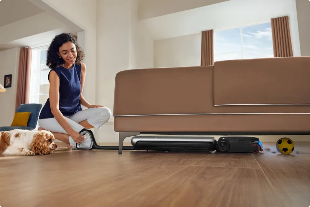 Dreame steam vacuum lying flat to reach under a low sofa while a dog rests nearby.