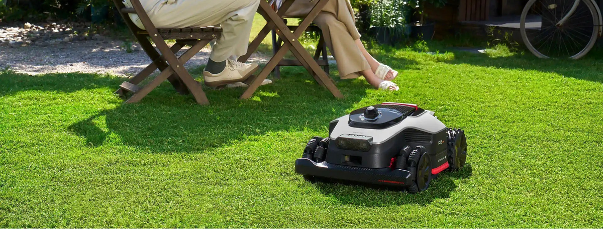 A couple plays chess at a garden table while a Dreame AI lawn mower cuts the grass behind them. 
