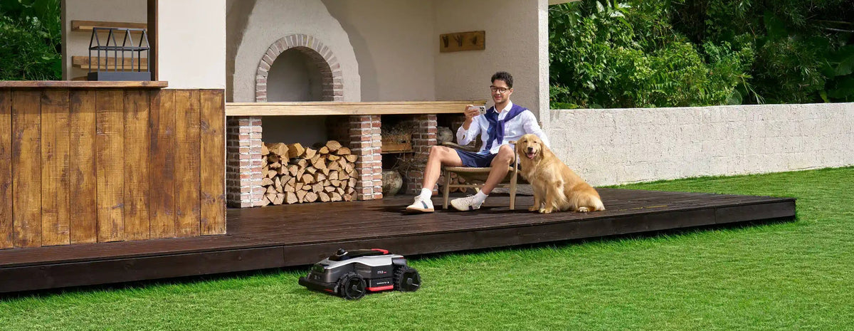Auto lawn mower cutting the backyard while a man and his pet relax on the patio