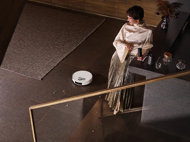 Dreame robot vacuum cleaning a dark tile floor with carpet and debris visible nearby, woman standing at a kitchen counter above.